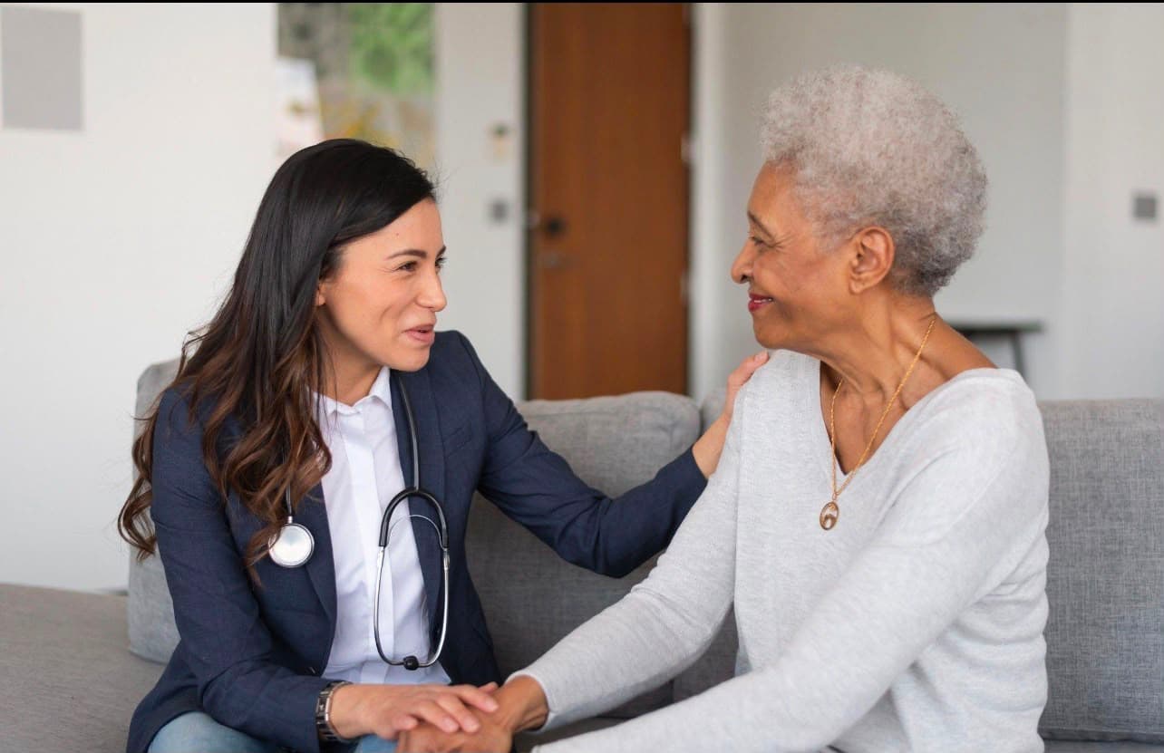 A clinician with a stethoscope sitting on a sofa with an older woman, holding her hand as they talk.