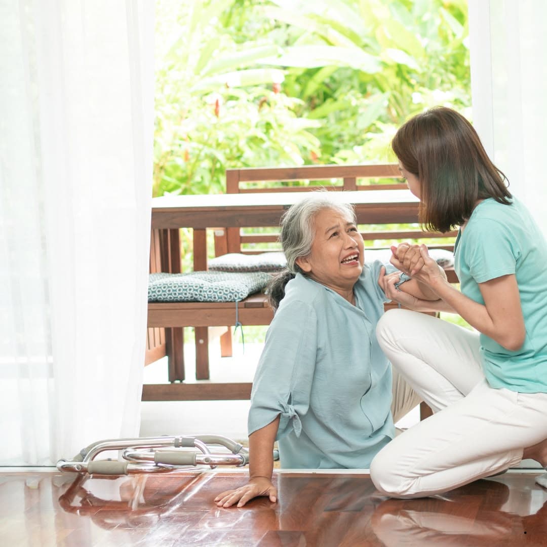 A woman kneeling beside an older woman who has fallen near a patio door, holding her hand and helping her up.