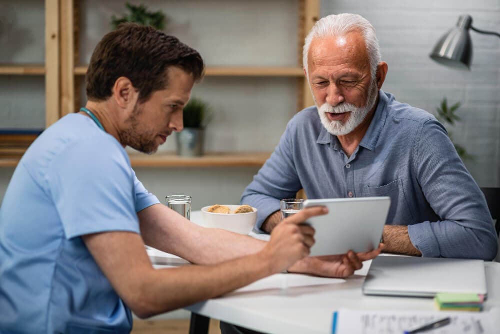 A care worker in scrubs sitting at a table, showing something on a tablet to an older man.