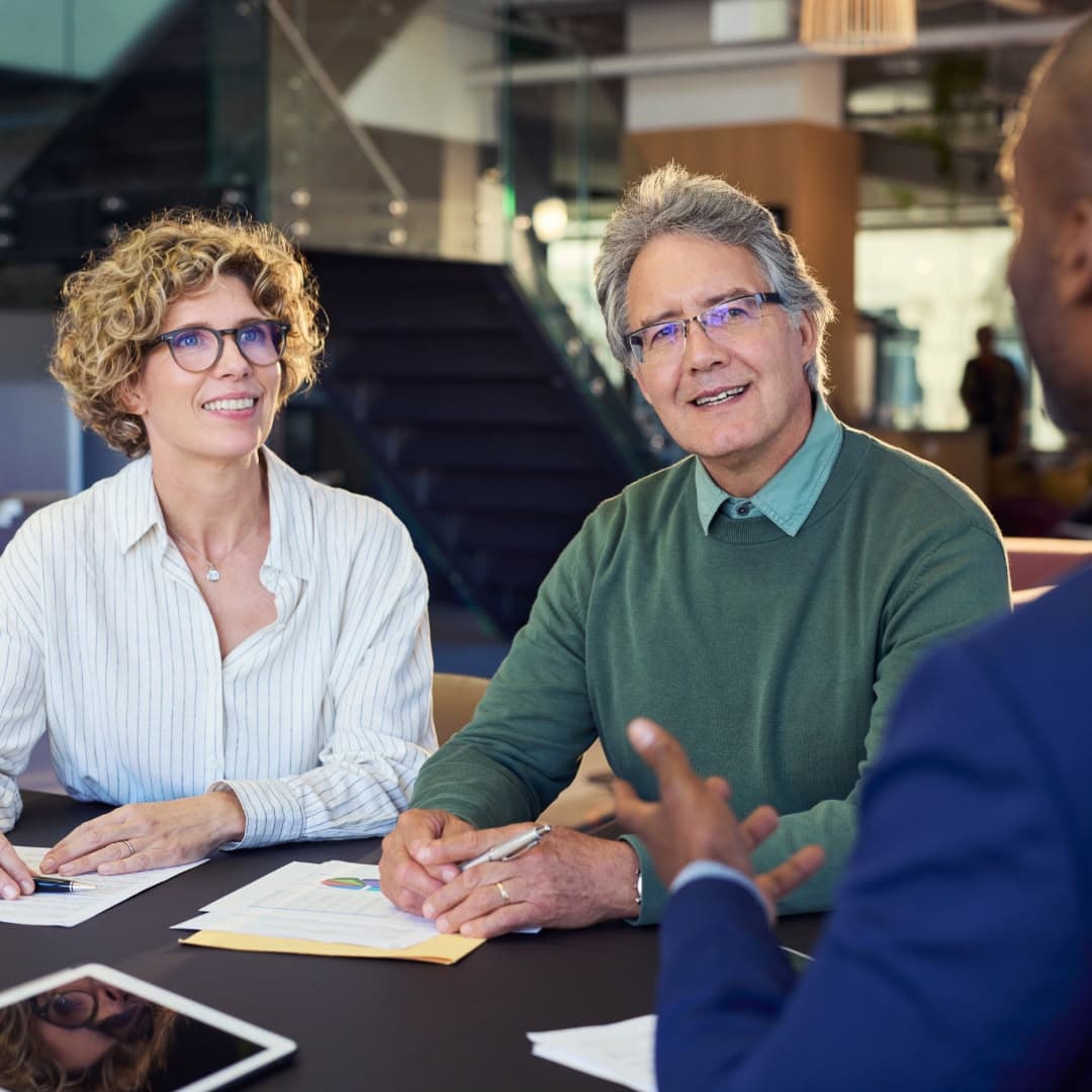 An older couple sitting at a table with paperwork, listening to an advisor across from them.
