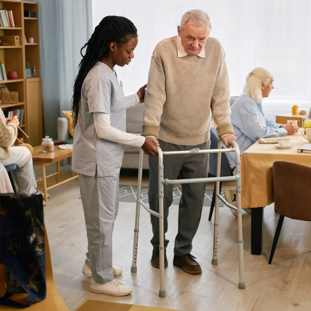 A care worker steadying an older man using a walker in a communal lounge.