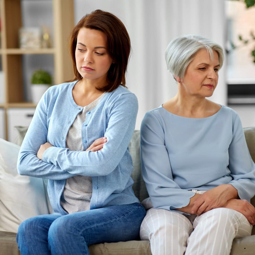 An adult daughter and her mother sitting on a sofa, both turned away from each other after a difficult conversation.
