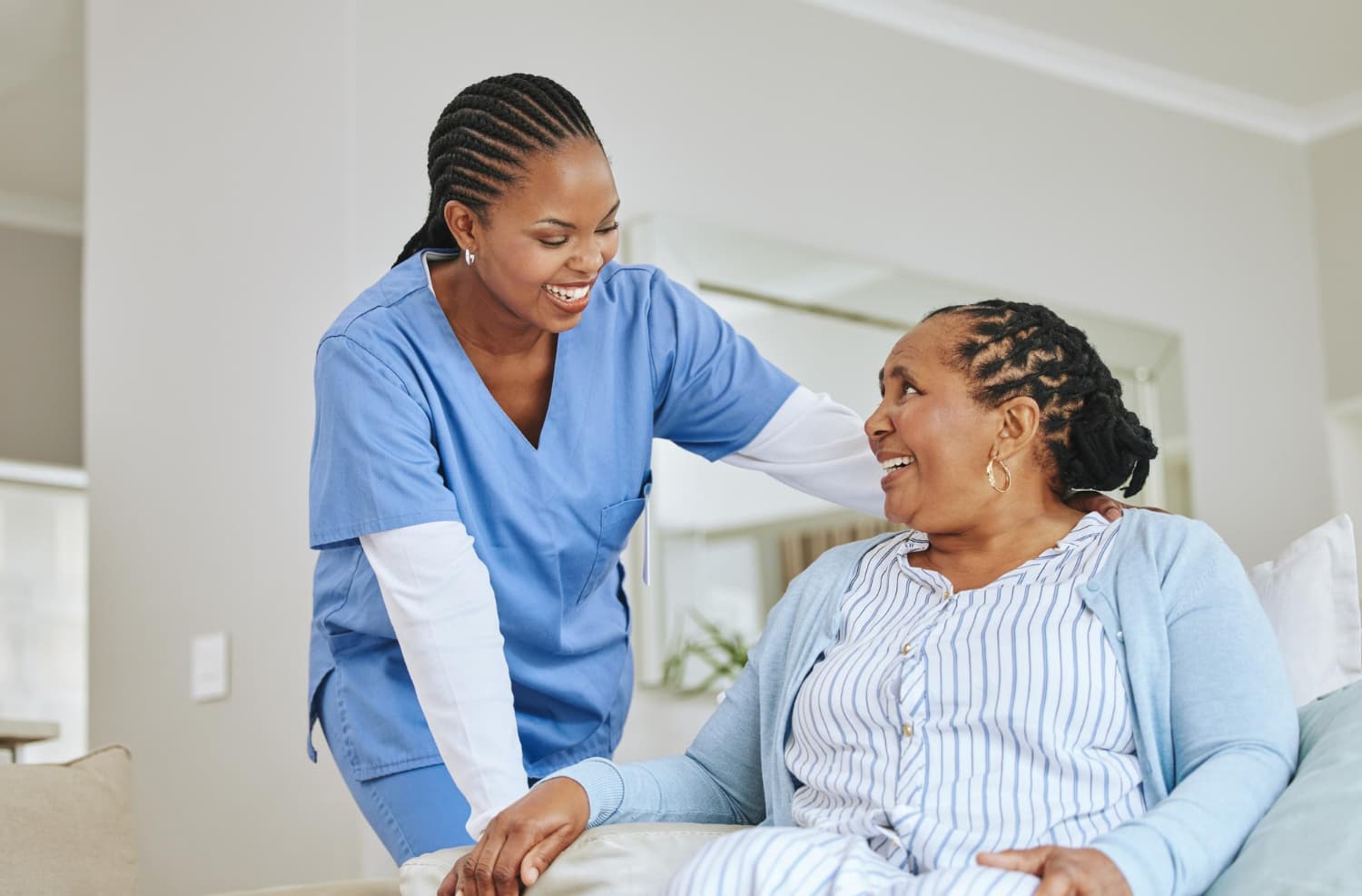 A nurse in scrubs leaning in to speak with a smiling older woman sitting up in bed.