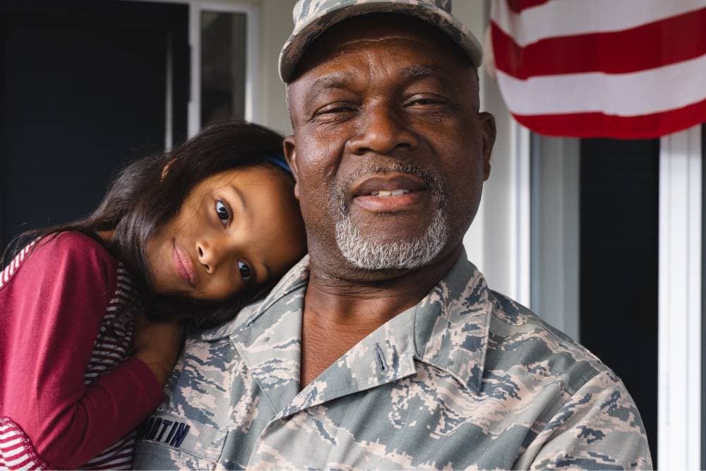 A young girl resting her head against a smiling veteran in uniform, a US flag behind them.