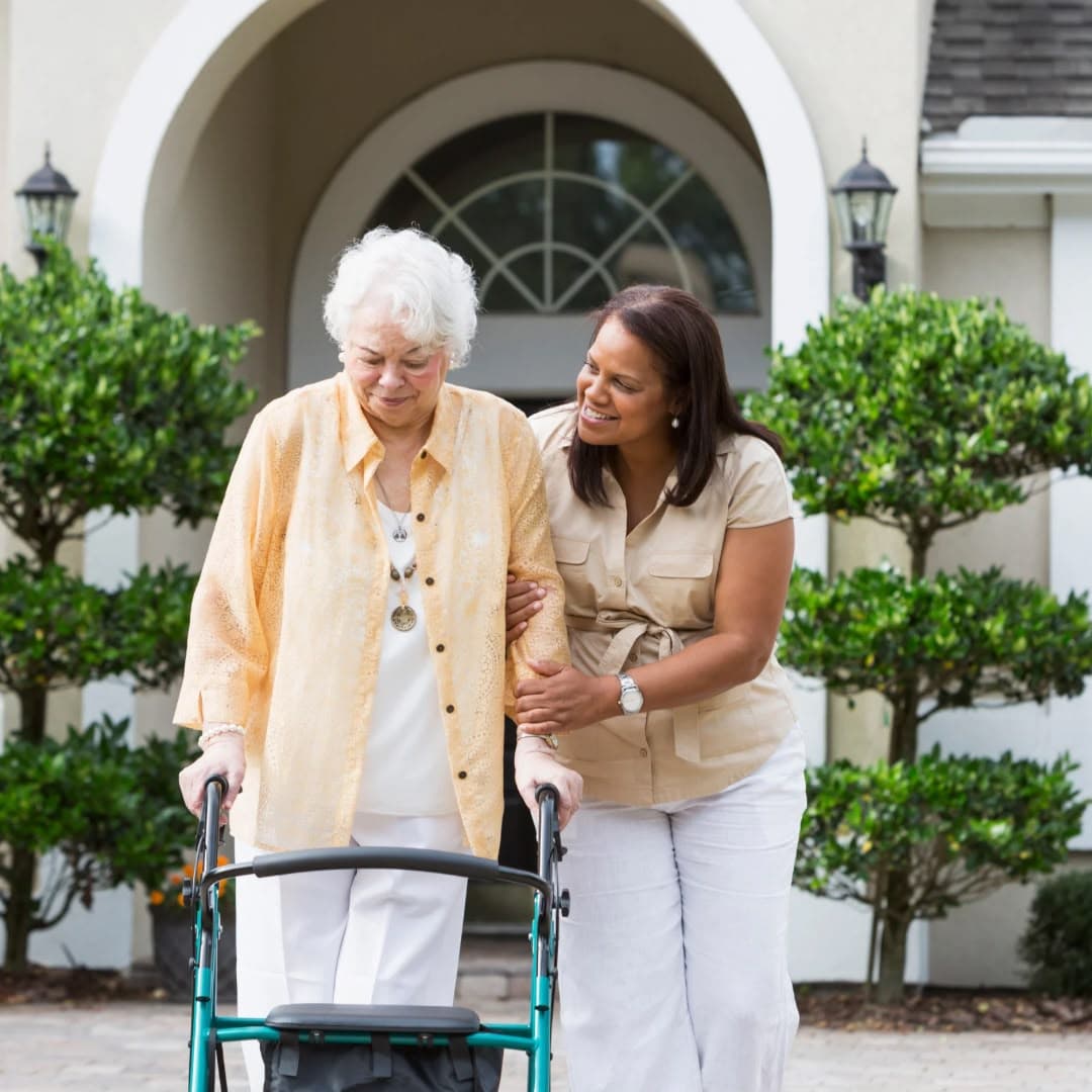 A woman walking beside an older woman using a wheeled walker along the front path of a house, steadying her arm.