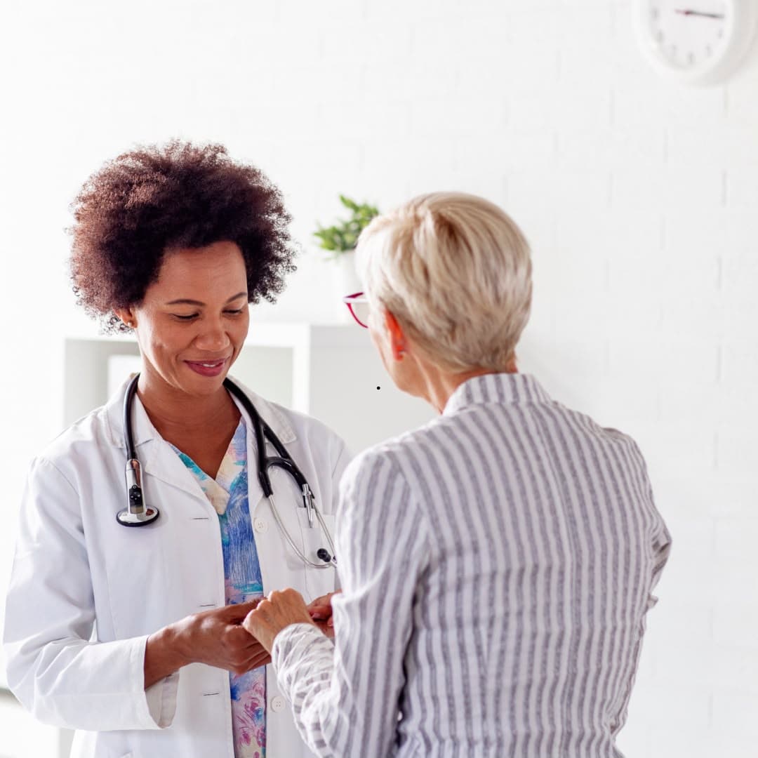 A doctor in a white coat holding an older woman's hands as the two of them talk in a bright consulting room.